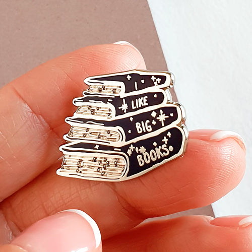 Pin de metal de 4 libros amontonados con la inscripción I like big books cogido en los dedos 